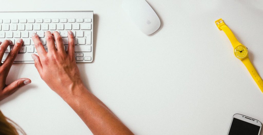 Hands typing on a keyboard next to a computer mouse and yellow watch